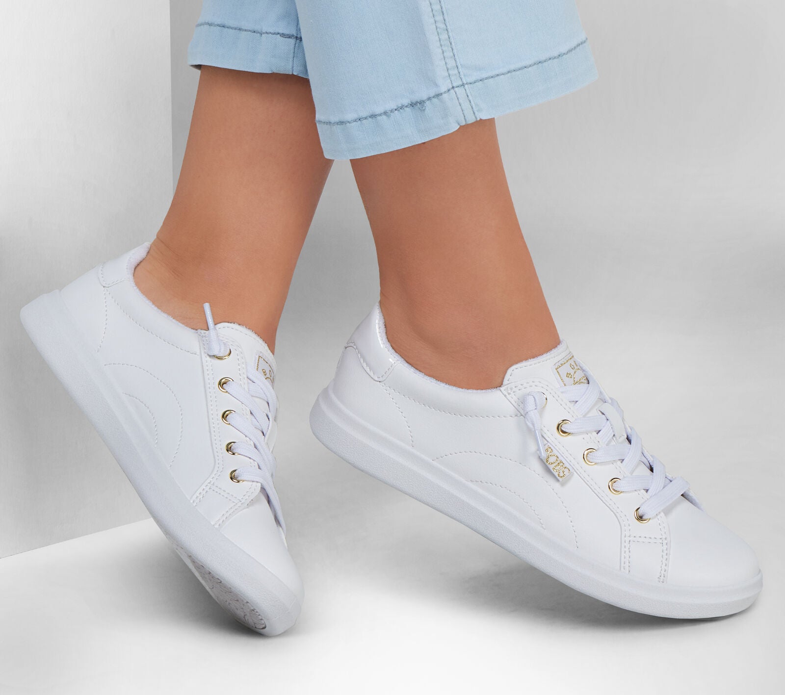 A pair of women's shoes by Skechers, style Bobs D'Vine 114453, in white canvas with rose gold accents and lace fastening. Lifestyle image.