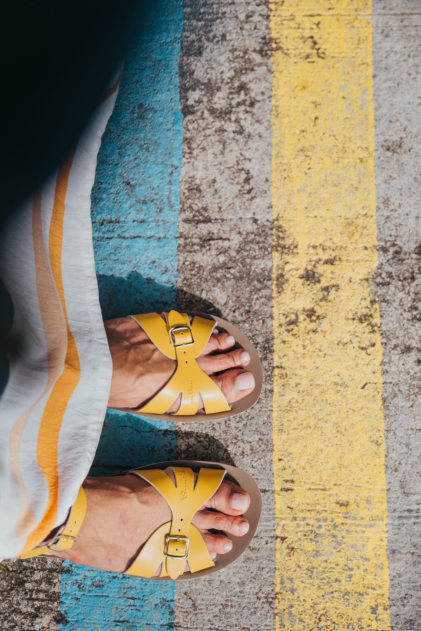 A Boardwalk style girls sandal by Salt Water Sandals in mustard with double buckle fastening across the toes and around the ankle. Open Toe and Sling-back. Lifestyle of looking down on a pair view.