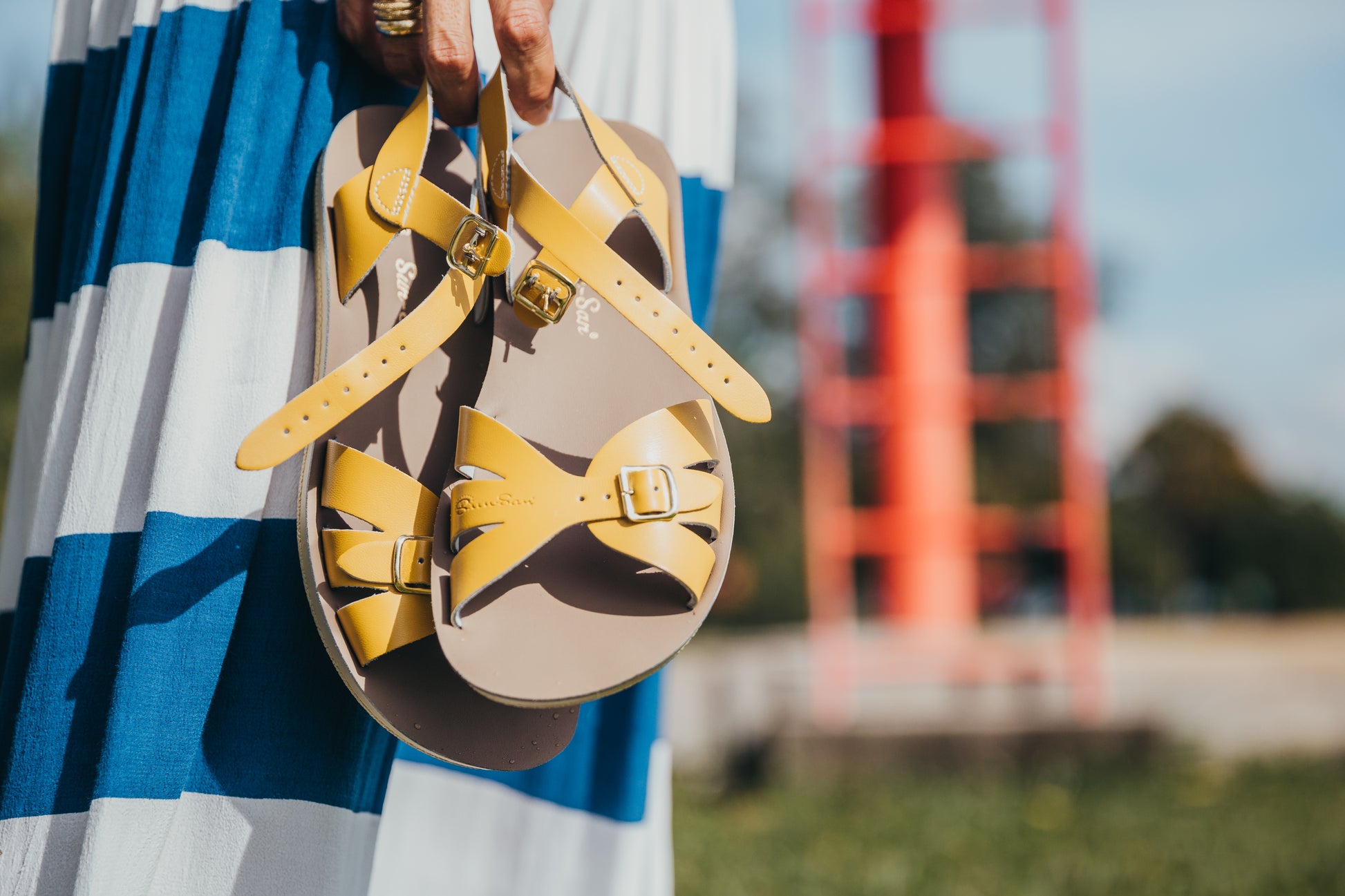 A Boardwalk style girls sandal by Salt Water Sandals in mustard with double buckle fastening across the toes and around the ankle. Open Toe and Sling-back. Lifestyle of lady holding a pair view.