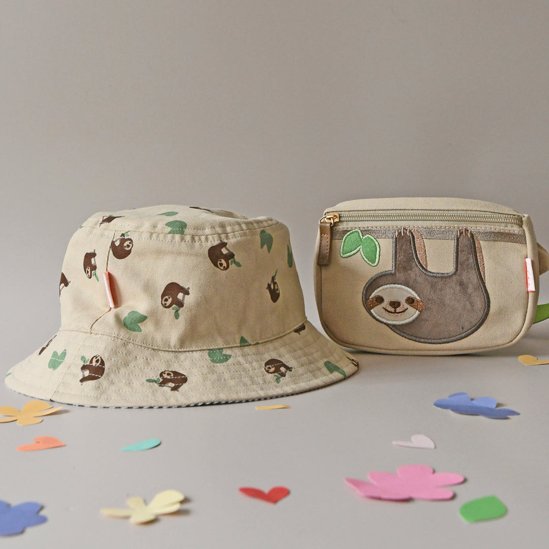 A pair of revirsable children's hats by Rockahula, style Sleepy Sloth, in beige with sleepy sloth print on one side and green stripe print on the other. Front view of hat with matching bum bag.