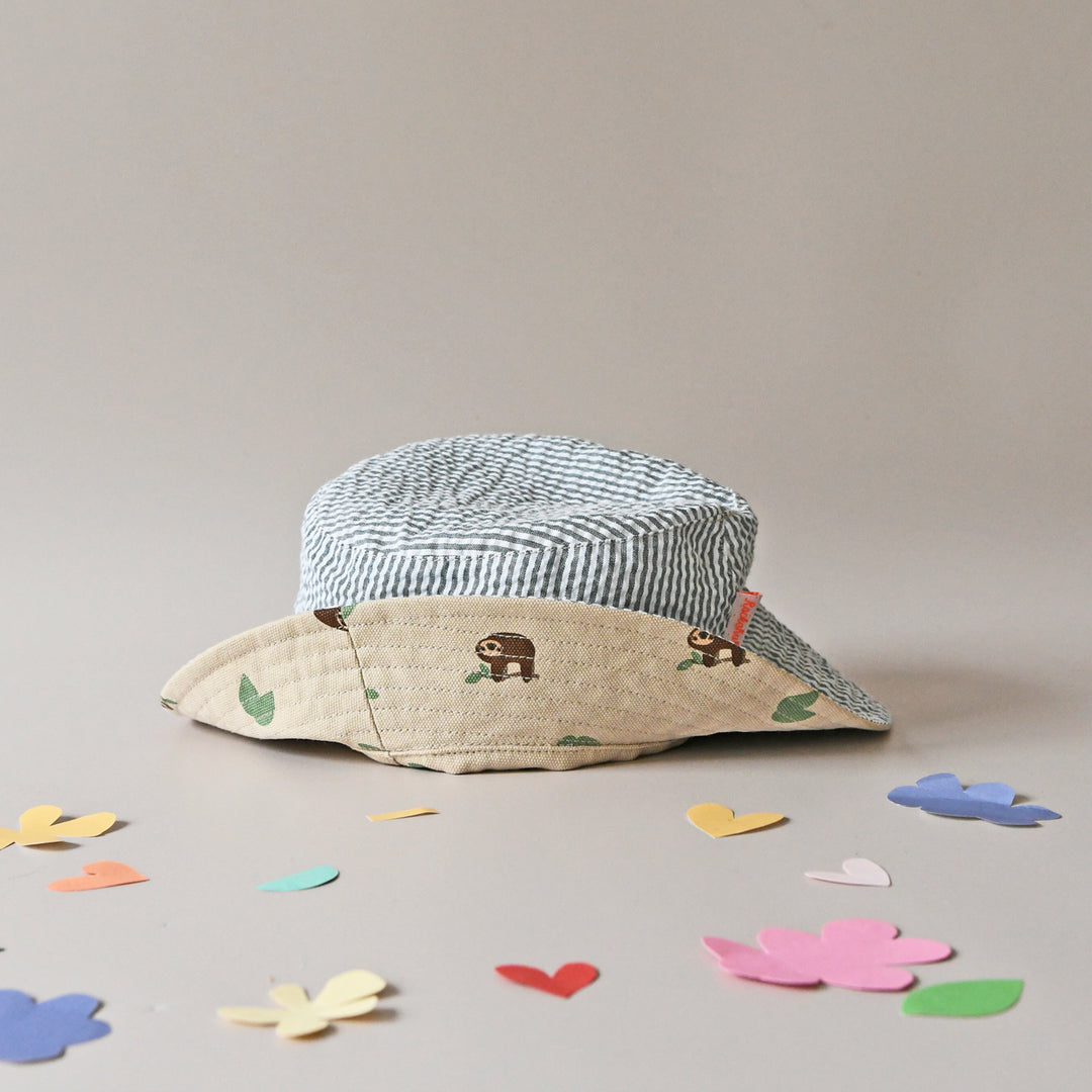 A pair of revirsable children's hats by Rockahula, style Sleepy Sloth, in beige with sleepy sloth print on one side and green stripe print on the other. Front view showing both sides of hat.