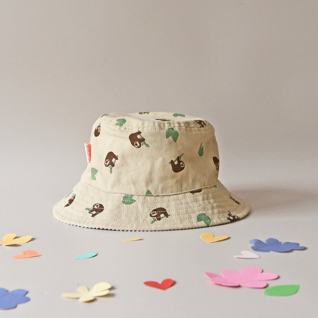 A revirsable children's hat by Rockahula, style Sleepy Sloth, in beige with sleepy sloth print on one side and green stripe print on the other. Front view of sleepy sloth side of hat.