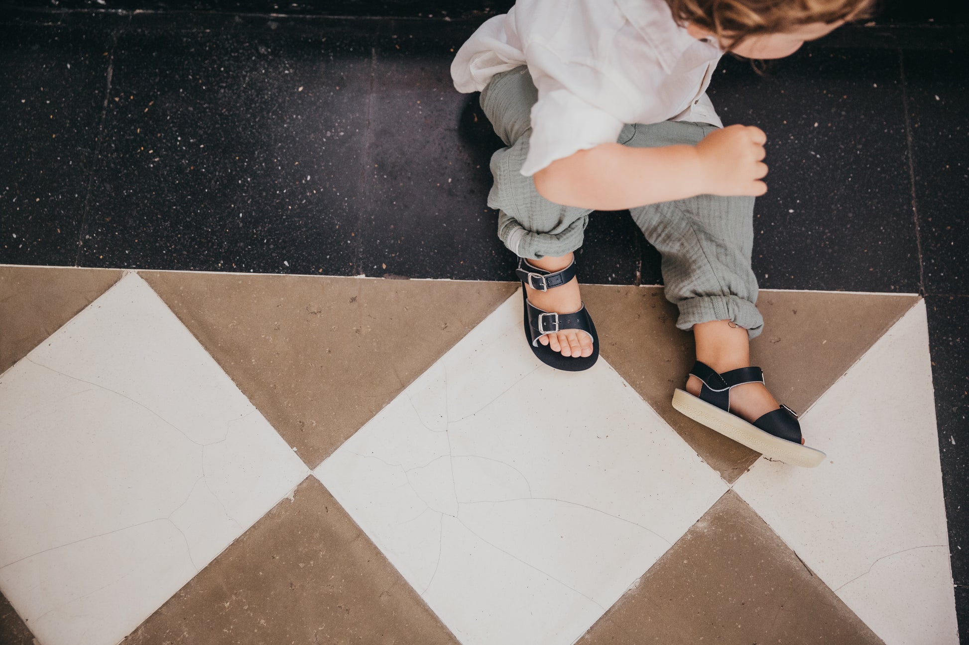 A unisex sandal by Salt Water Sandals in navy with double buckle fastening across the instep and around the ankle. Open Toe and Sling-back. Lifestyle of boy sat on floor.