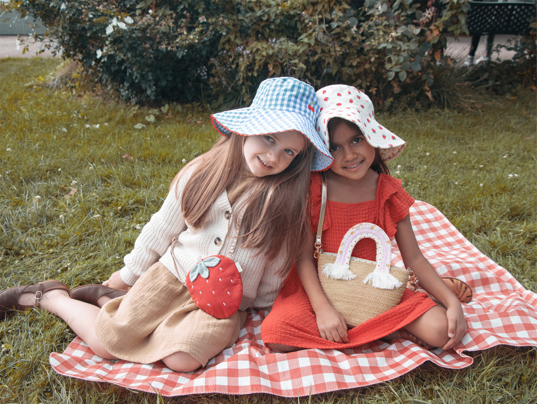 A reversible girls bucket hat by Rockahula, style Cherry Gingham,one side is blue and white stripe cherry print, the other blue and white gingham with cherry embroidery. Lifestyle image showing gingham side of hat.