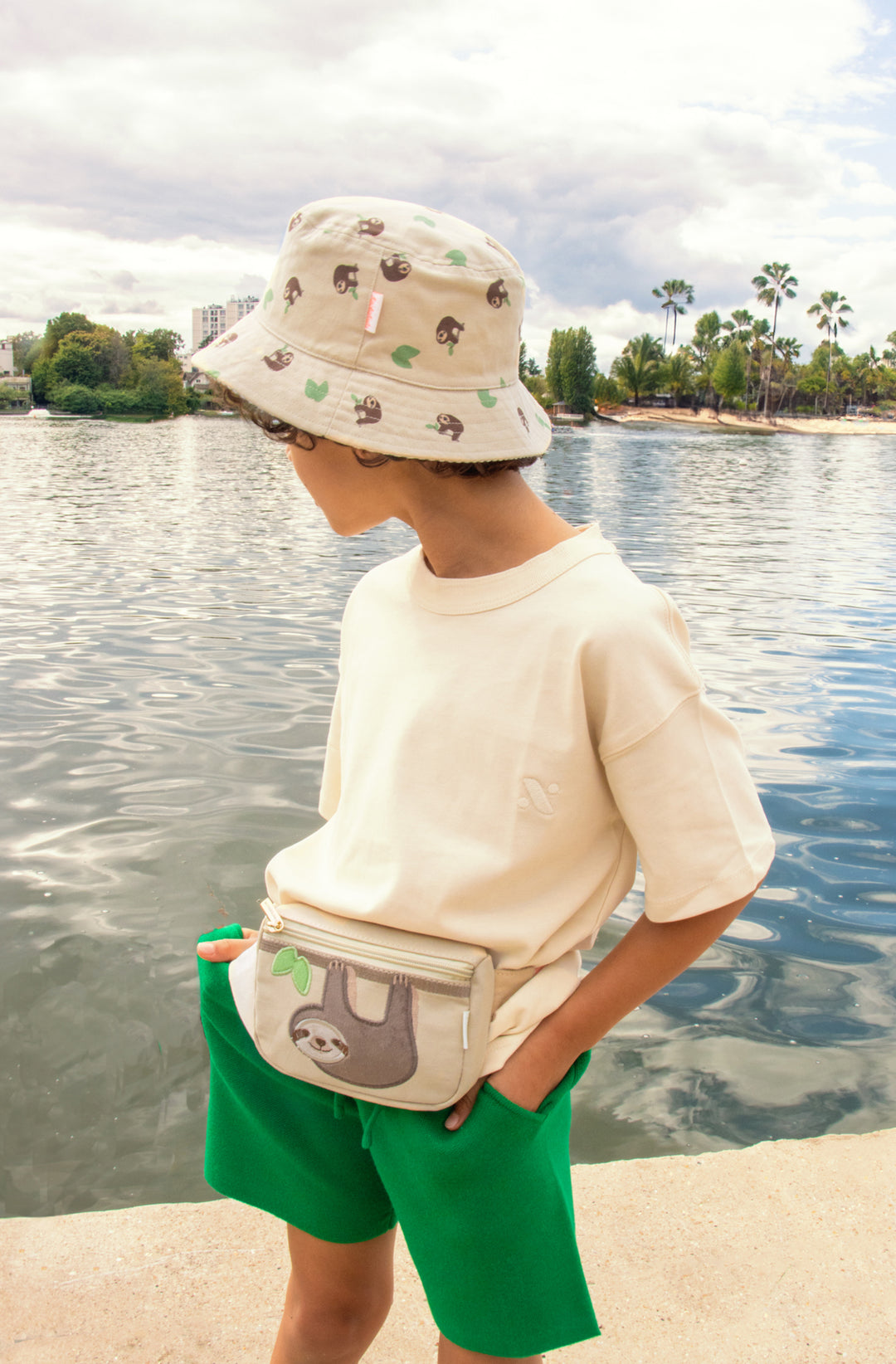 A pair of revirsable children's hats by Rockahula, style Sleepy Sloth, in beige with sleepy sloth print on one side and green stripe print on the other. Lifestyle image.