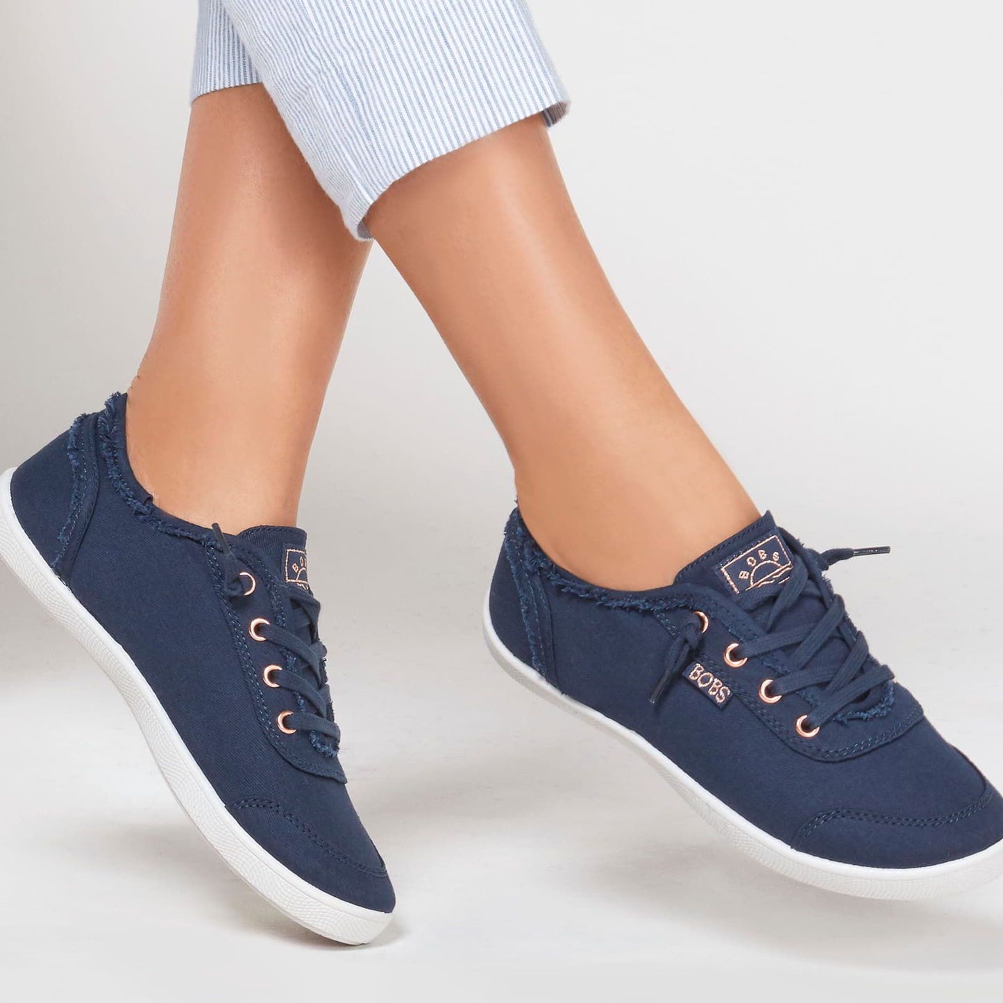 A pair of women's shoes by Skechers, style Bobs D'Vine 114453, in navy canvas with rose gold accents and lace fastening. Lifestyle image.